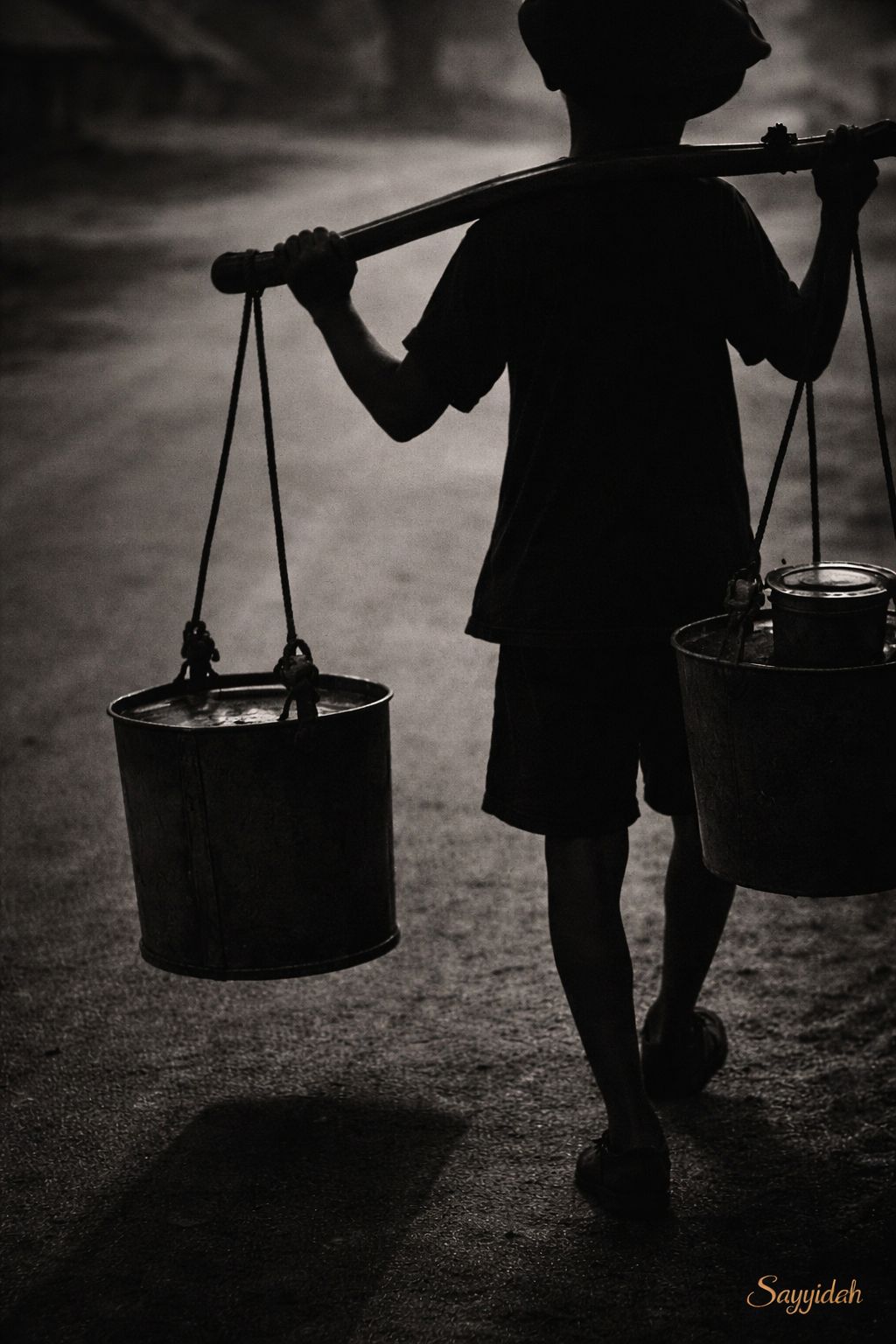 Black and white silhouette of a person carrying water buckets along a quiet road at dawn in fine art style